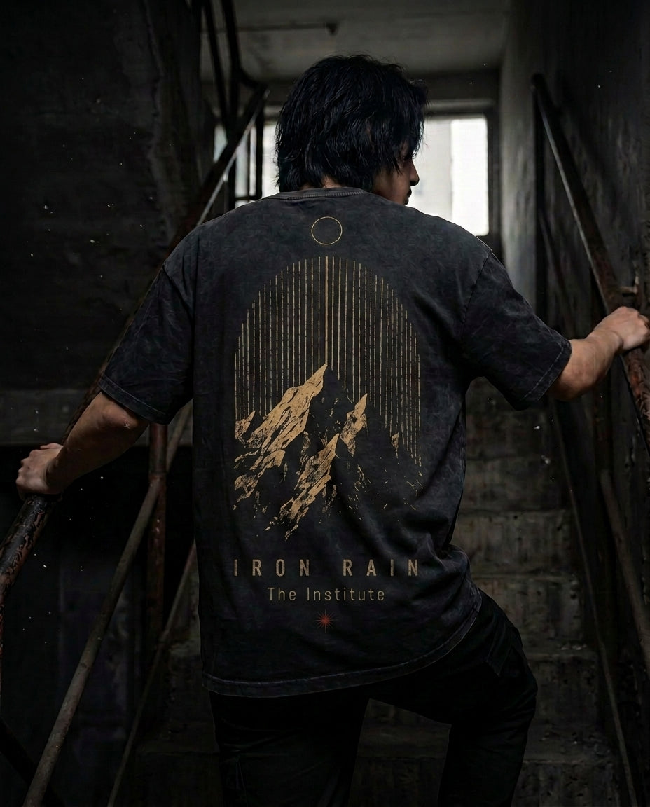 A man viewed from behind wearing a washed black t-shirt with a large gold graphic of mountains and the text 'IRON RAIN The Institute' on the back, standing on a dark concrete staircase.